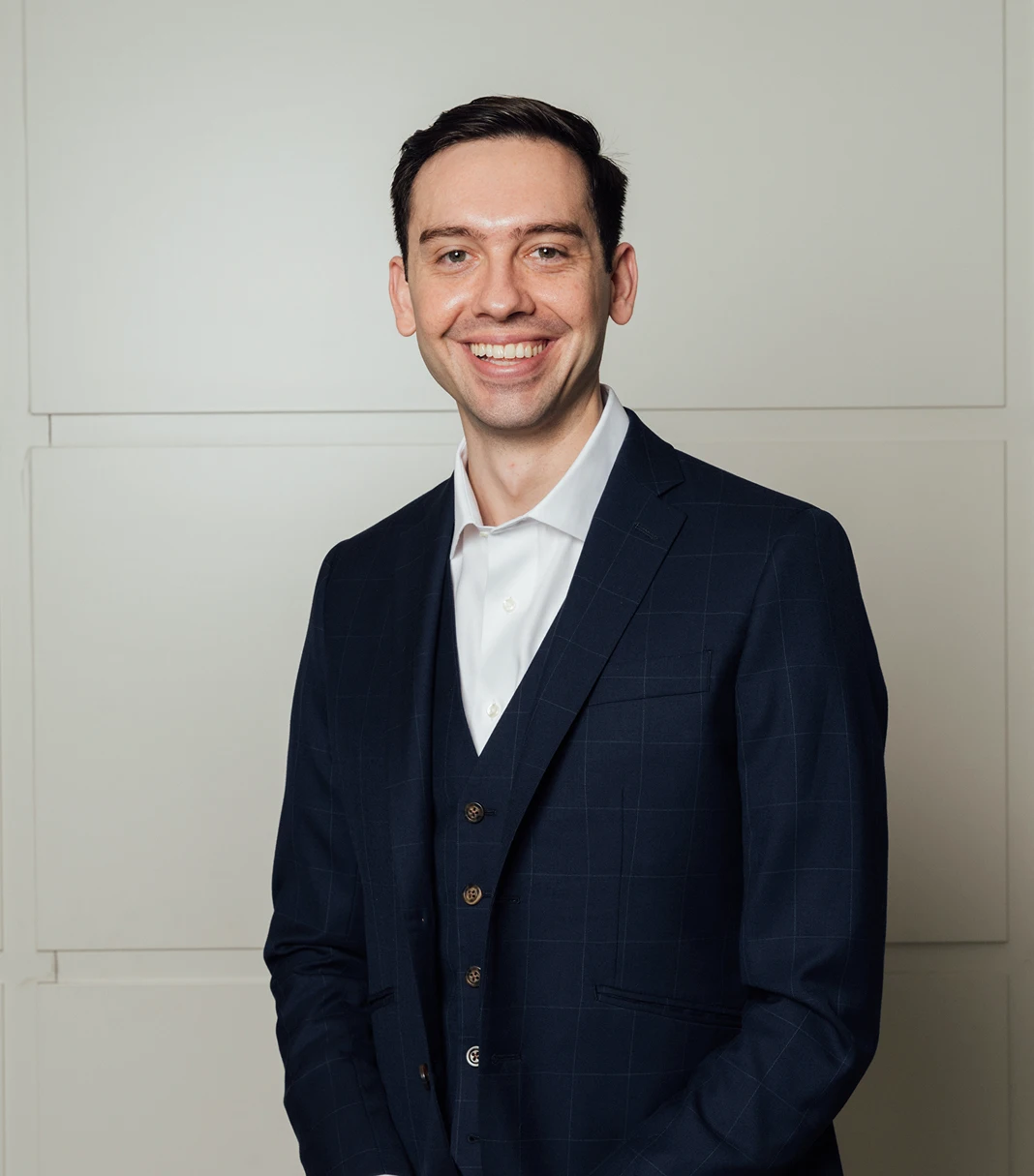 A professional waist-up portrait of Dr. Evan Jones, who is smiling warmly at the camera. He is wearing a sophisticated navy blue three-piece windowpane suit over a crisp white dress shirt. He stands against a minimalist, light grey paneled wall that provides a clean and modern backdrop. - Doctor in Los Angeles, CA