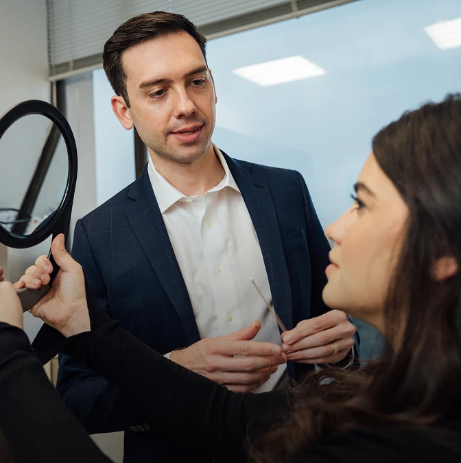 A male clinician in a dark suit and white shirt standing in a bright office, holding a medical swab while consulting with a woman who is looking into a handheld mirror. - Face in Los Angeles, CA