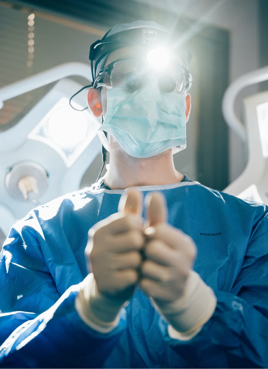 A surgeon in blue scrubs, a surgical mask, and a lighted headlamp stands in an operating room giving a double thumbs-up.- Facelift in Los Angeles, CA