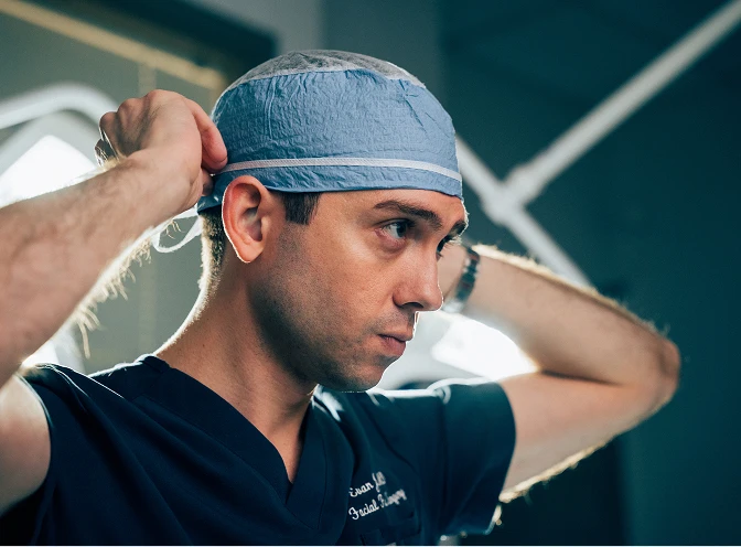 A side-profile shot of a male surgeon in dark blue scrubs, focused and preparing for a procedure. He is shown from the chest up, reaching back with both hands to tie a light blue surgical cap. His expression is serious and concentrated. The background is a dimly lit clinical environment with a bright, blurred light source providing a sharp rim light along his features. - About in Los Angeles, CA
