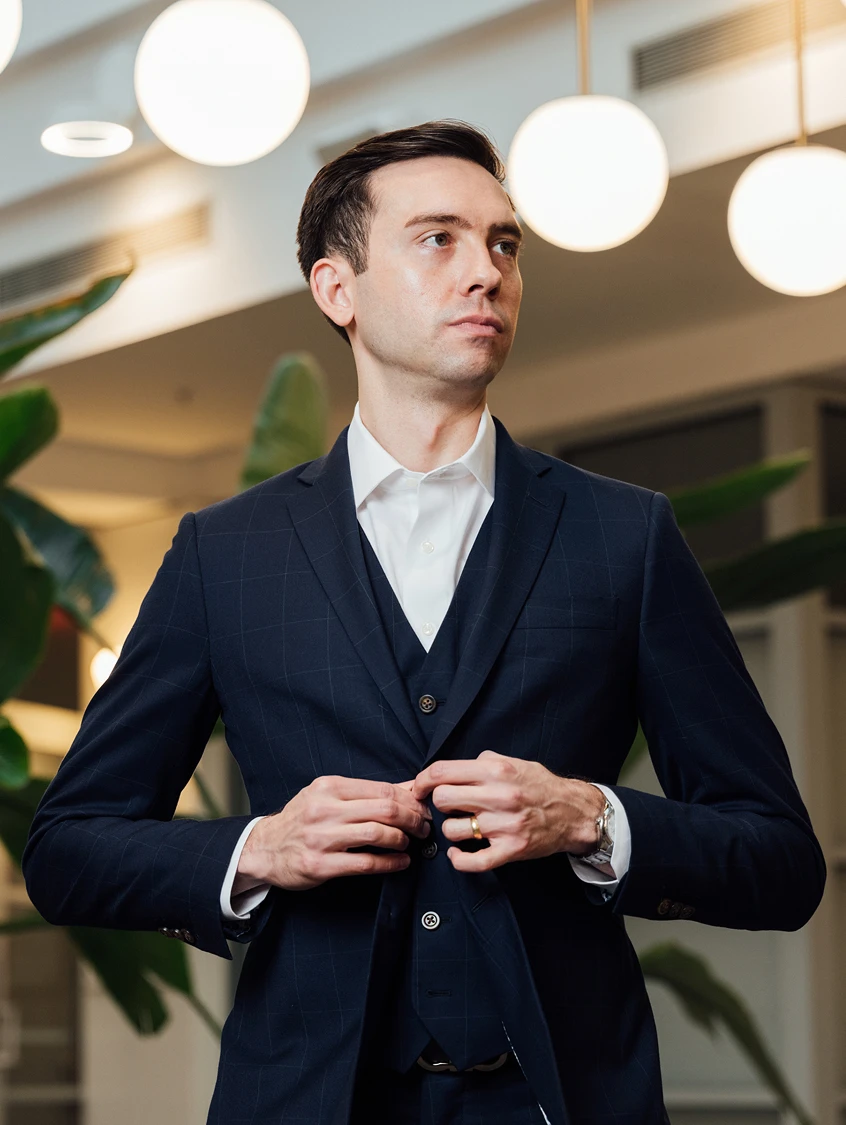 A low-angle, professional portrait of Dr. Evan Jones wearing a sophisticated navy blue three-piece windowpane suit over a crisp white shirt. He is captured in a mid-action pose, buttoning his blazer while looking off-camera with a composed and serious expression. The background is a modern, high-ceilinged interior featuring large spherical pendant lights and lush green indoor plants, creating an elegant and professional ambiance. - About in Los Angeles, CA