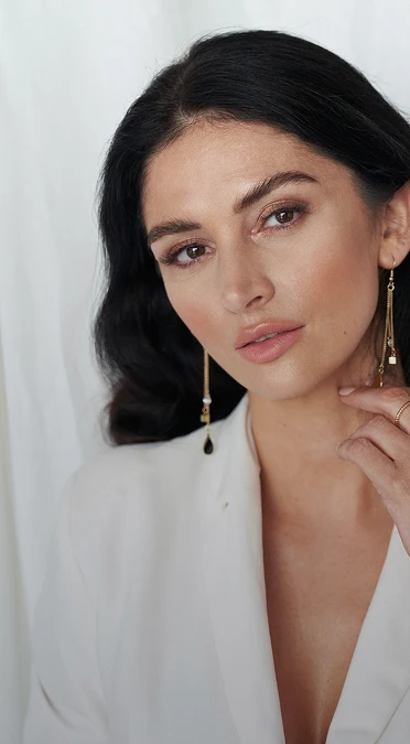 A vertical portrait of a woman with dark wavy hair and a polished makeup look, wearing a white blazer. She is looking directly at the camera with a neutral expression, resting her hand lightly against her neck. She wears long, gold-toned drop earrings with geometric charms and pearls. The background is a soft, draped white fabric that creates a clean, high-fashion aesthetic. - Brow lift in Los Angeles, CA