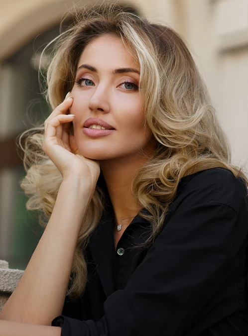 A portrait of a woman with voluminous blonde hair and soft pink makeup, wearing a black button-down shirt. She is leaning forward with her chin resting on her hand, looking toward the camera with a subtle, confident expression. The outdoor background is softly blurred, showing the neutral tones of a building in natural light. - Buccal in Los Angeles, CA