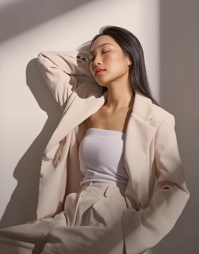 A portrait of a woman with long dark hair, wearing a cream-colored blazer and matching trousers over a white tube top. She is leaning against a white wall with her eyes closed and one arm raised behind her head, bathed in warm, direct sunlight that creates sharp shadows. The aesthetic is minimalist and serene. - Laser in Los Angeles, CA