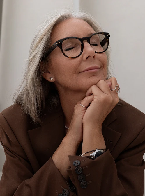 A portrait of a mature woman with stylish grey hair and tortoiseshell glasses, wearing a chocolate brown blazer. She has her eyes closed and a peaceful, slight smile on her face, with her hands clasped under her chin. She is wearing several rings and a delicate silver watch. The background is a minimalist, off-white wall, giving the image a serene and sophisticated feel. - Home in Los Angeles, CA