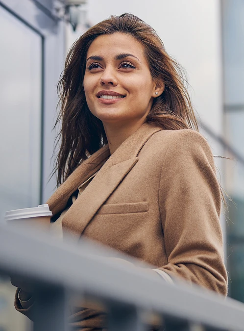 A medium outdoor shot of a smiling woman with long brown hair, wearing a tan wool blazer. She is looking off-camera with a confident expression while holding a disposable coffee cup. The background features the clean, blurred lines of a modern glass building, creating a professional and bright urban atmosphere. - Scar in Los Angeles, CA