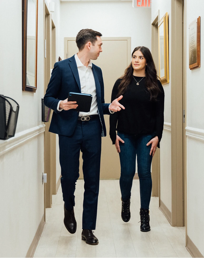 A male clinician in a blue suit and a female patient in a black sweater and jeans walk together down a brightly lit medical office hallway while engaged in conversation. - first step in Los Angeles, CA
