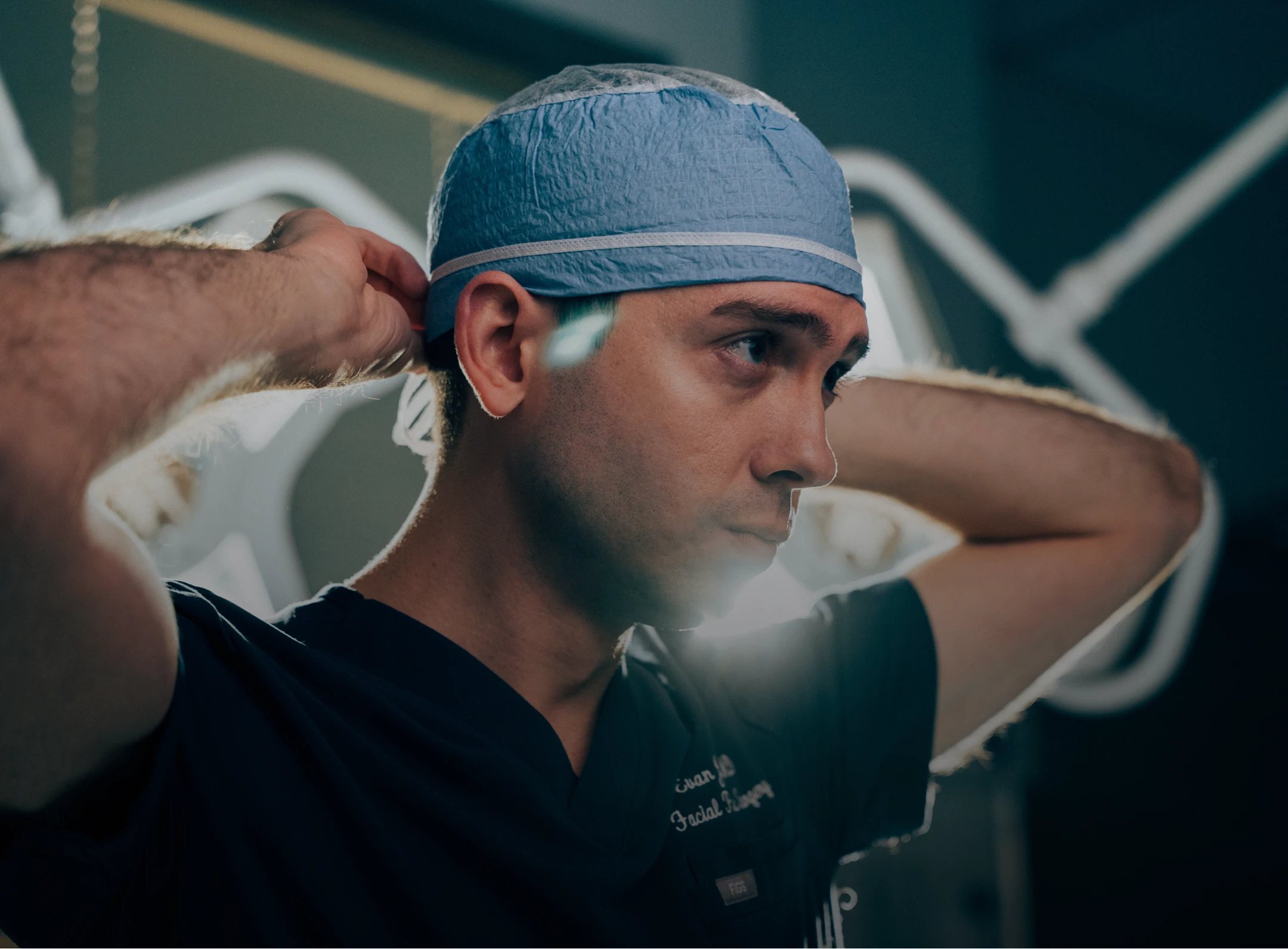 A surgeon in dark scrubs ties a blue surgical cap behind his head while standing in a dimly lit operating room under bright surgical lights. - Procedure in Los Angeles, CA
