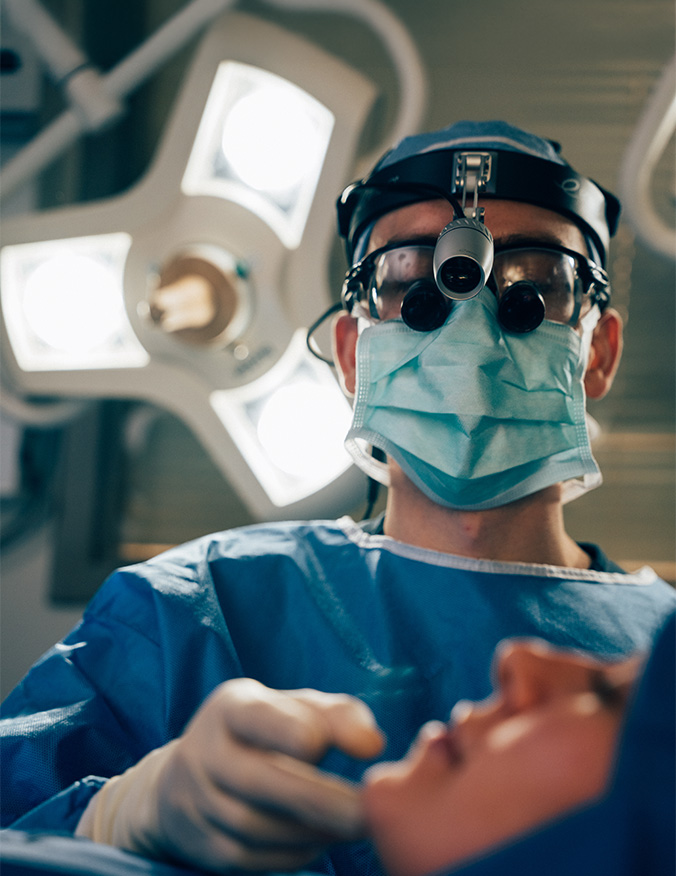 A close-up, low-angle photograph from an operating room showing a surgeon in blue scrubs, a mask, and specialized surgical loupes with a mounted headlamp. The surgeon is looking down toward a patient, whose chin and mouth are visible in the soft-focus foreground. In the blurred background, a large, multi-bulb surgical light provides a bright, clinical glow that illuminates the sterile environment. - Chin augmentation in Los Angeles, CA