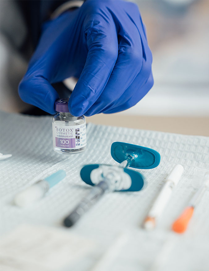 A close-up shot of a hand in a bright blue surgical glove holding a small, clear glass vial labeled "BOTOX COSMETIC." The vial is positioned above a sterile white medical tray containing various treatment supplies, including a syringe with blue finger grips and several capped needles. The focus is sharp on the vial and the gloved fingertips, with the background softly blurred to emphasize the medical preparation. - Botox and Wrinkle relaxers in Los Angeles, CA