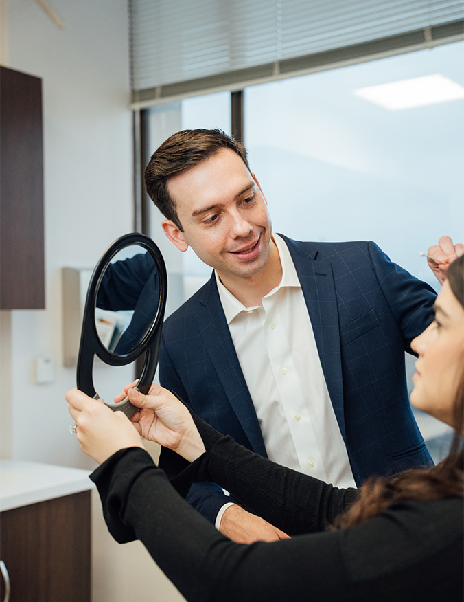 A professional consultation in a clinical setting where a male specialist in a navy blazer and white shirt observes a female patient. The patient holds a round black hand mirror, looking at her reflection while the specialist points a cotton swab toward her brow area. The room is bright and modern, featuring clean white walls and dark wood cabinetry in the background. - Brow lift in Los Angeles, CA