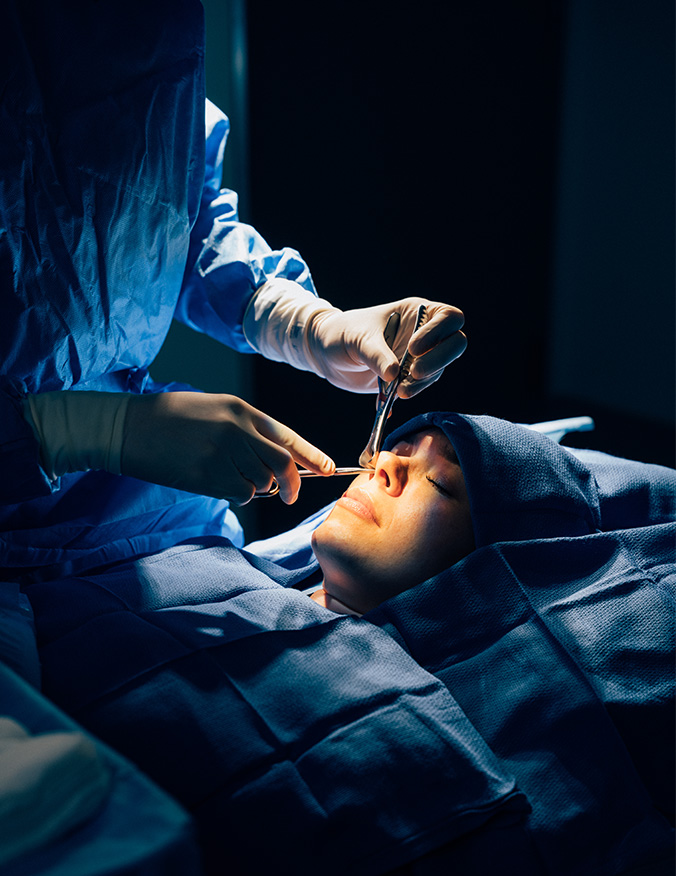 A dramatic, low-angle shot of a surgeon in a sterile operating room, seen from the perspective of the patient. The surgeon wears blue scrubs, a mask, and surgical loupes with a bright headlamp, looking down with focus as they begin a procedure. - cosmetic rhinoplasty in Los Angeles, CA