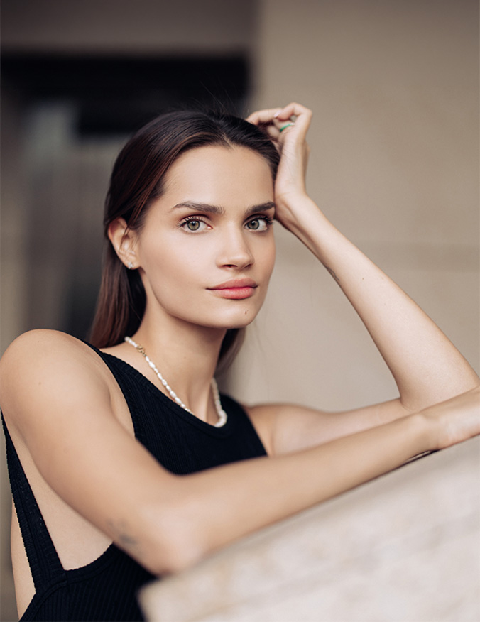 A woman with her arms resting on a slanted flat surface. She wears a black tank top and pearl necklace. She has long brown hair that is straight and behind her back. She has light eyes and looks directly at the camera. - Ear surgery and earlobe repair in Los Angeles, CA