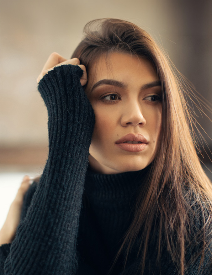 A portrait of a woman with long, dark hair wearing a black ribbed turtleneck. She is looking off-camera with a thoughtful expression, resting one hand against her head, which highlights her smooth forehead and the area around her eyes. - Eyelid lift in Los Angeles, CA