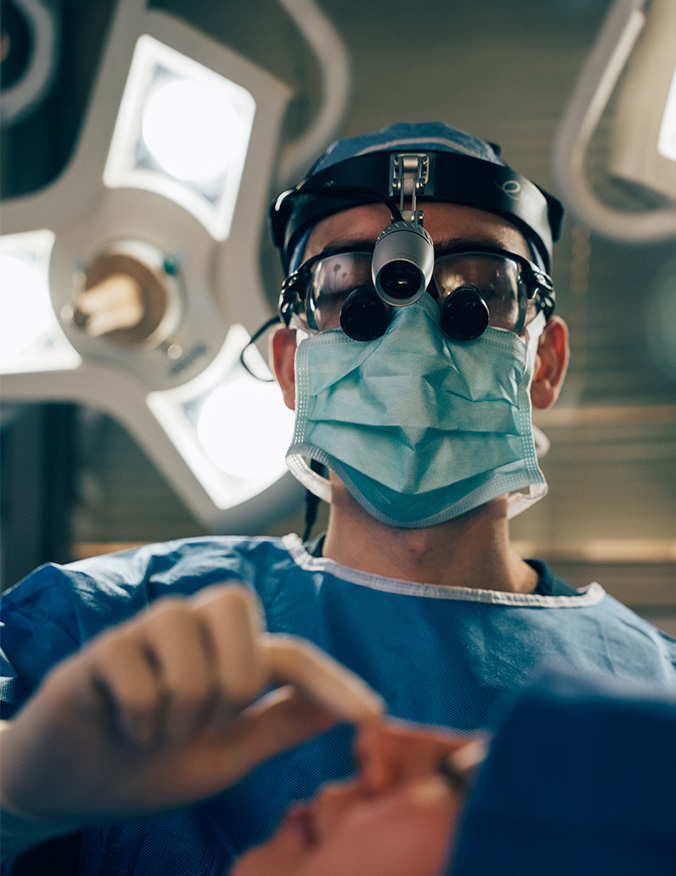 A dramatic, low-angle shot of a surgeon in a sterile operating room, seen from the perspective of the patient. The surgeon wears blue scrubs, a mask, and surgical loupes with a bright headlamp, looking down with focus as they begin a procedure. - Functional rhino on Los Angeles, CA