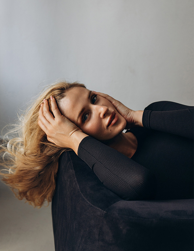 A woman with blonde hair lying back on a dark, plush surface. She is looking directly at the camera with her hands gently framing her forehead and temples, emphasizing her clear skin and facial features against a neutral background. - Laser skin resurfacing in Los Angeles, CA