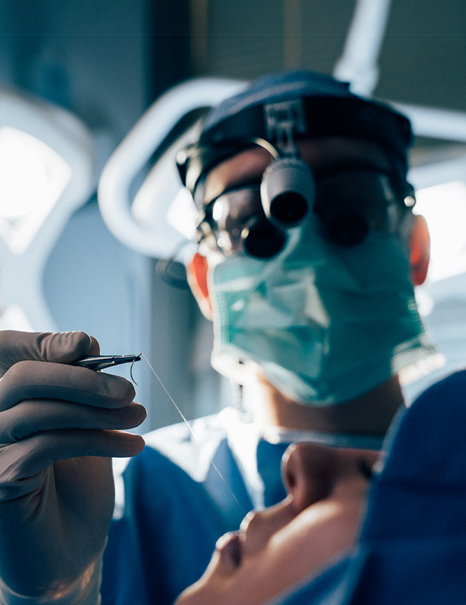 A close-up, low-angle shot from the patient's perspective during surgery. A surgeon wearing a mask, cap, and surgical loupes holds a fine needle and suture thread, focused on the patient's face which is partially visible in the foreground. - Lip lift in Los Angeles, CA