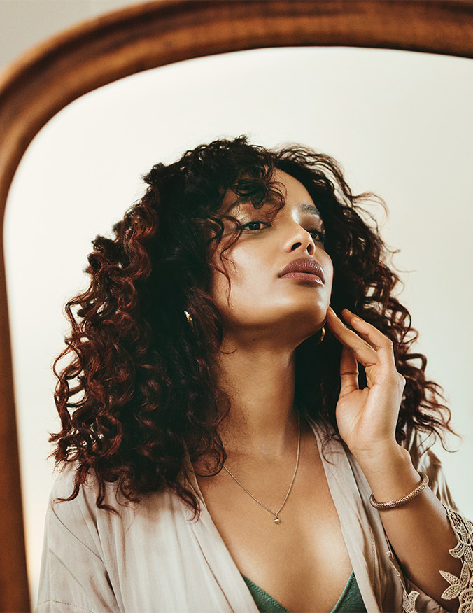 A portrait of a woman with voluminous, dark curly hair looking upward and slightly away from the camera. She is gently touching her jawline with her fingers, showcasing her neck profile and defined chin against a bright, airy background. - Neck and chin liposuction in Los Angeles, CA