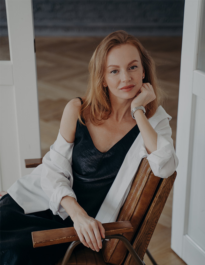 A woman with strawberry blonde hair sitting in a leather chair, looking directly at the camera with a subtle smile. She is leaning her head on her hand, highlighting a smooth neck and jawline while wearing a black top and an open white button-down shirt. - Neck lift in Los Angeles, CA