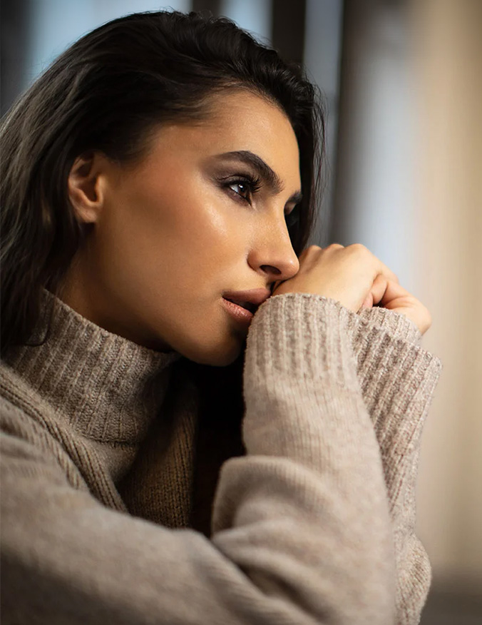 A profile portrait of a woman with long dark hair wearing a beige turtleneck sweater. She is resting her chin on her hands and looking thoughtfully into the distance, showcasing her straight nasal bridge and refined facial profile in soft, natural lighting. - Revision rhino in Los Angeles, CA