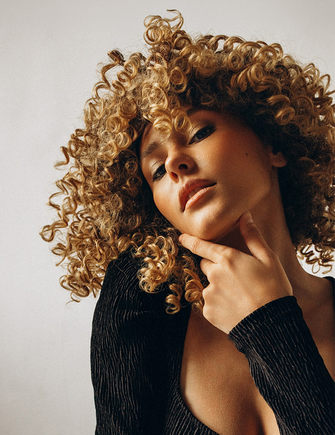 A portrait of a woman with voluminous, tight golden curls leaning her head back slightly. She has her hand resting under her chin, highlighting her jawline and smooth skin texture, set against a plain, light-colored background. - Scar revision in Los Angeles, CA
