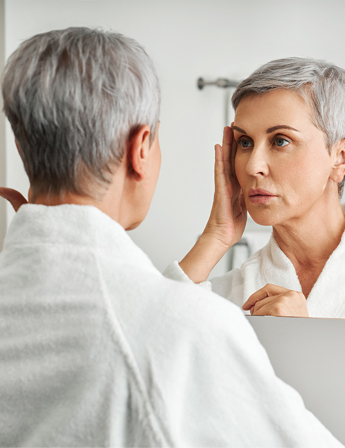 A woman with short gray hair, seen from behind in a white robe, looking at her reflection in a bathroom mirror. She is gently touching her temple and examining her facial skin with a serious, observant expression. - Skin cancer reconstruction MOHS in Los Angeles, CA