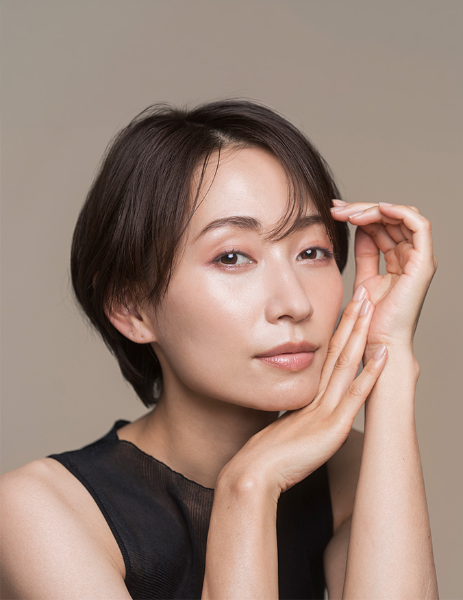 A portrait of a woman with short dark hair and a clear complexion, looking directly at the camera with a neutral expression. She has her hands gently framing her face, highlighting her cheekbones and smooth skin against a soft, tan background. - Structural fat grafting in Los Angeles, CA