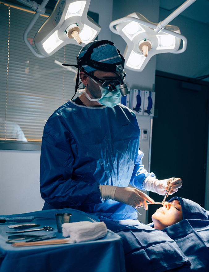A wide vertical shot of a surgeon in a sterile operating room, wearing blue scrubs, a surgical mask, and a head-mounted magnifying light. He is focused on performing a rhinoplasty on a patient whose face is partially visible under surgical drapes, with bright overhead surgical lights illuminating the workspace. - Tip rhinoplasty in Los Angeles, CA